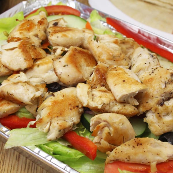 Grilled chicken pieces on a bed of fresh lettuce, tomatoes, cucumbers, and olives in a foil container.