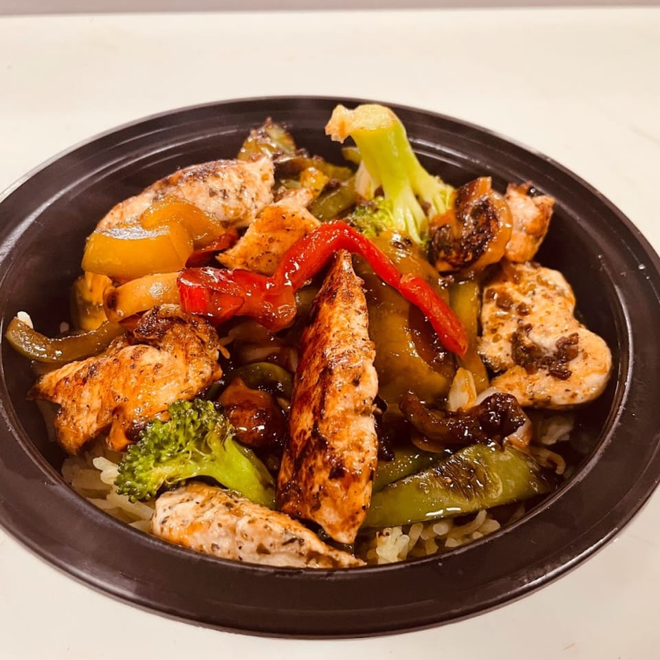 A close-up of a chicken and vegetable stir-fry with broccoli, peppers, and onions over rice in a black bowl.